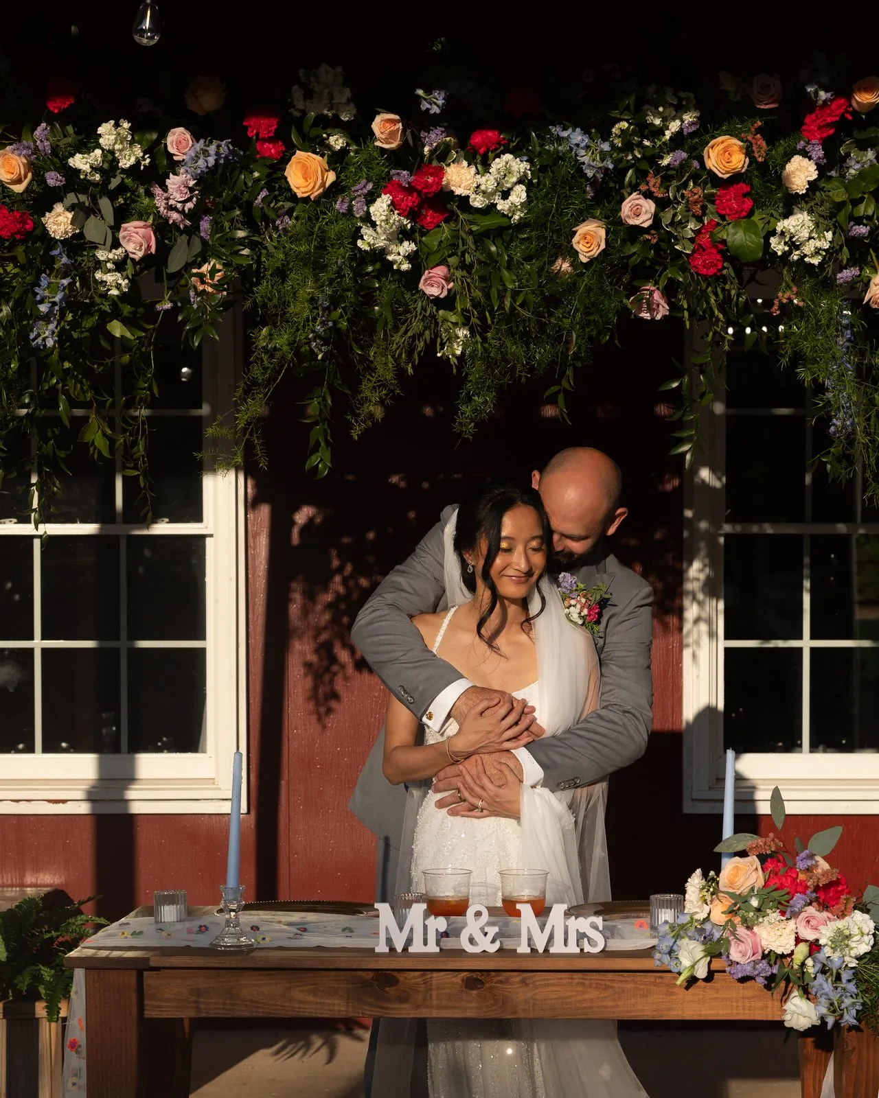 Couple at their sweetheart table beneath a lush floral garland at The Heritage Barn